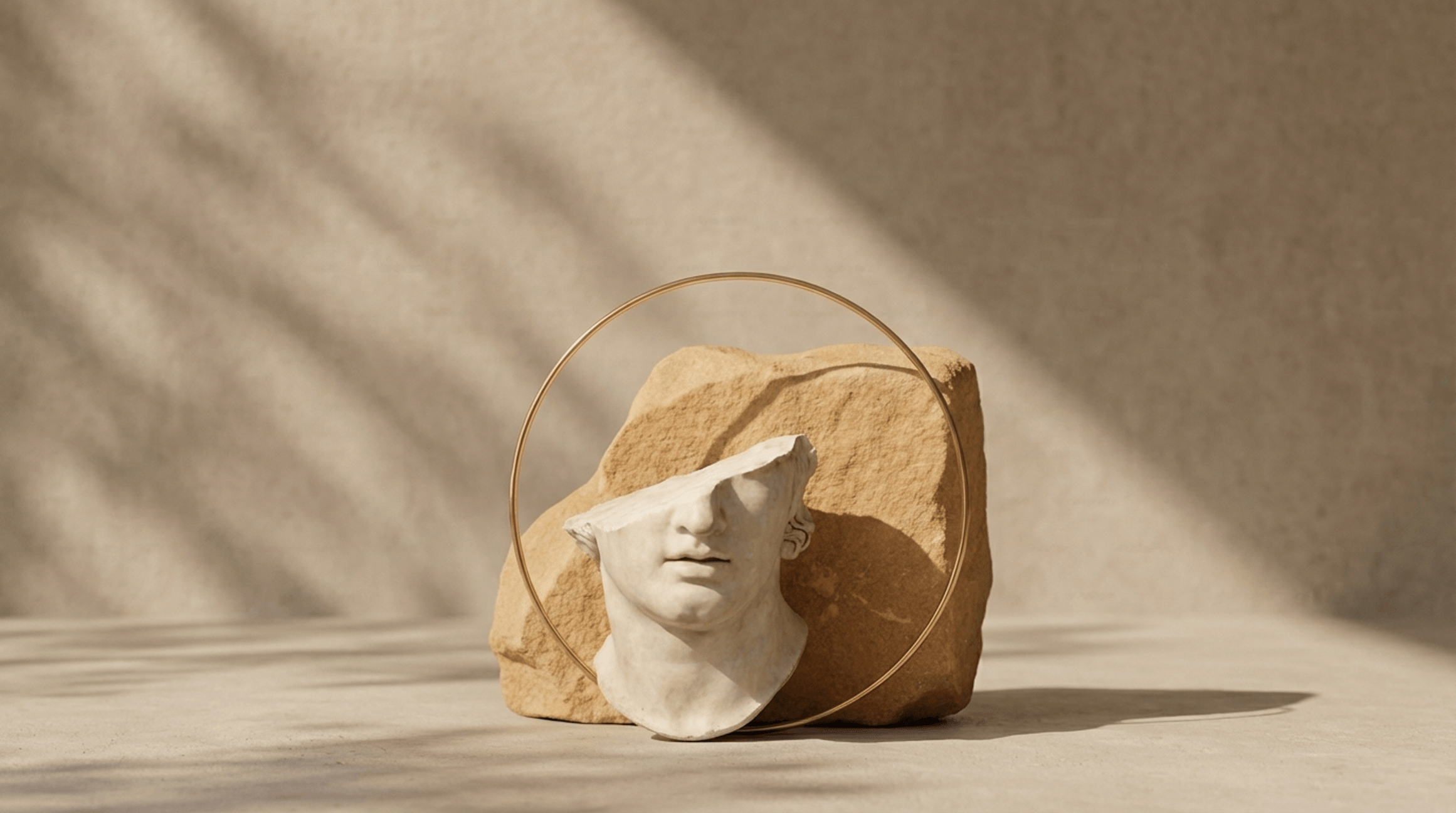 A broken white classical bust stands against a large textured tan rock, encircled by a gold hoop, on a sunlit beige surface with strong shadows.