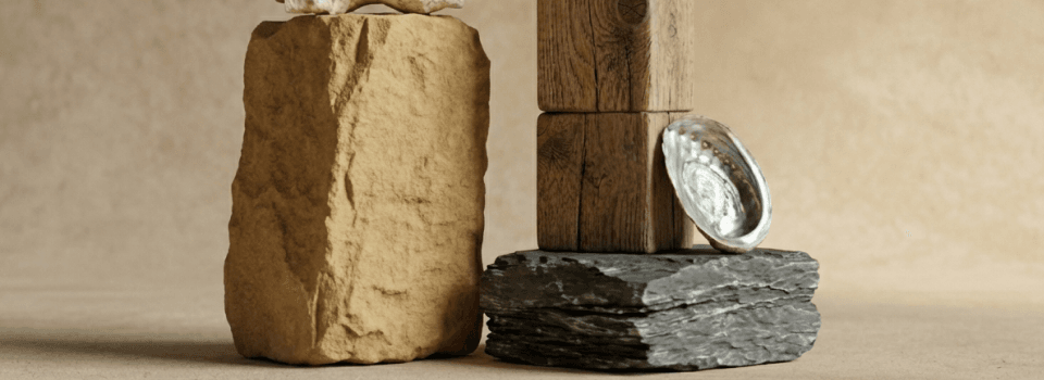A display of natural elements featuring a tall, rough sandstone block, stacked weathered wooden blocks, layered dark slate, and an iridescent abalone shell.